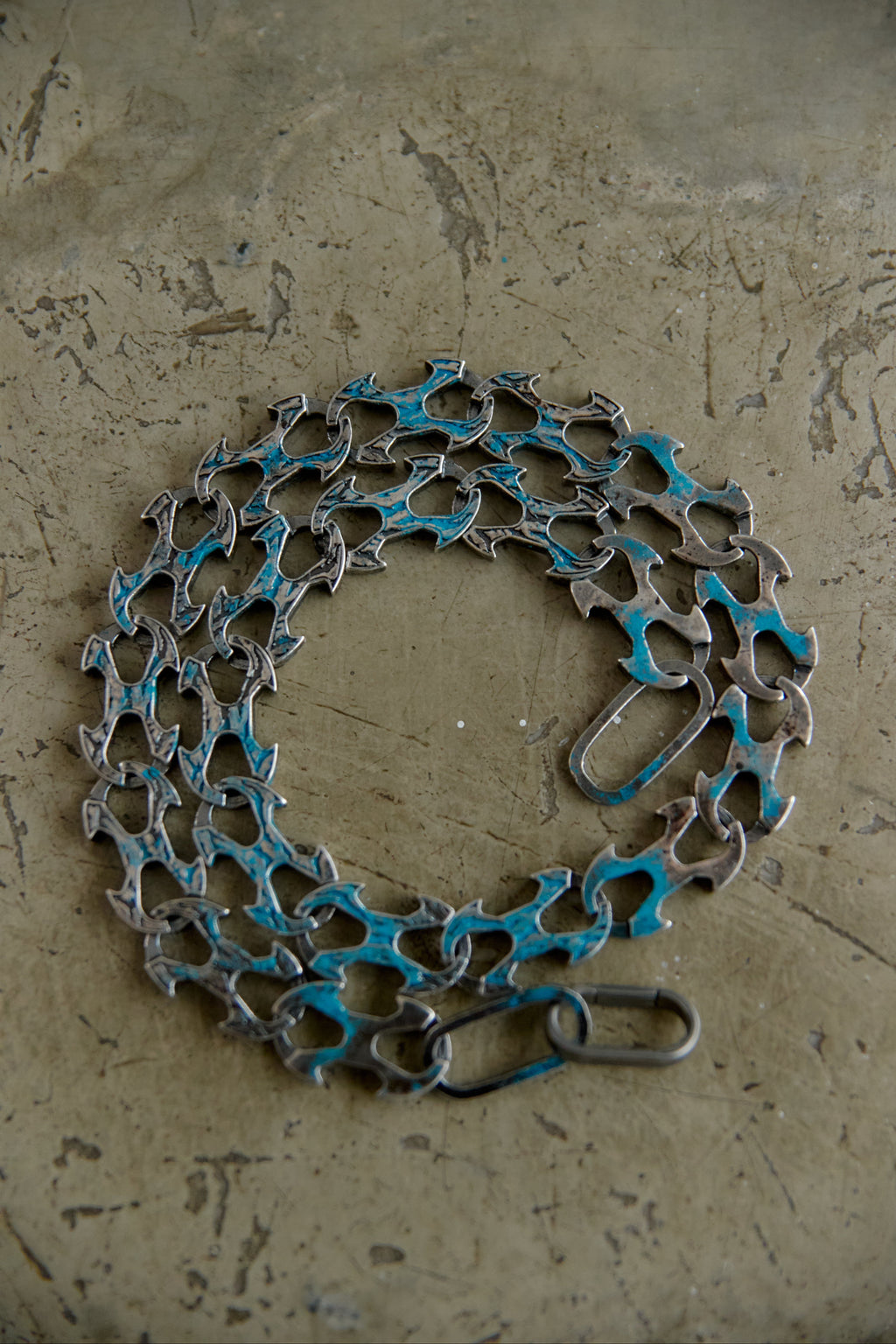 A bicycle chain wrapped in blue painterly material and secured with a metal clasp, displayed on a textured concrete surface.