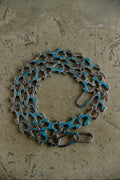 A bicycle chain wrapped in blue painterly material and secured with a metal clasp, displayed on a textured concrete surface.