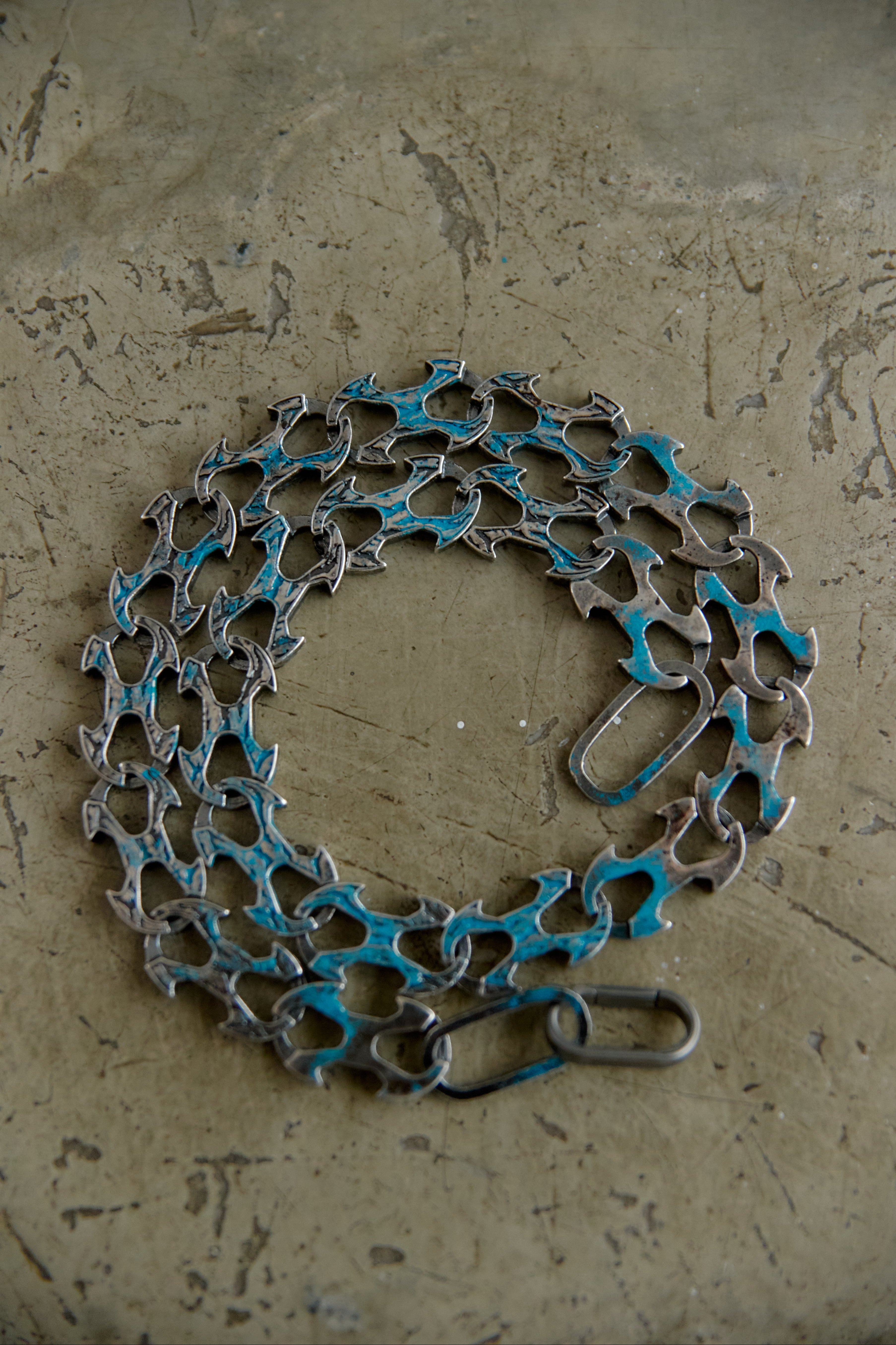 A bicycle chain wrapped in blue painterly material and secured with a metal clasp, displayed on a textured concrete surface.