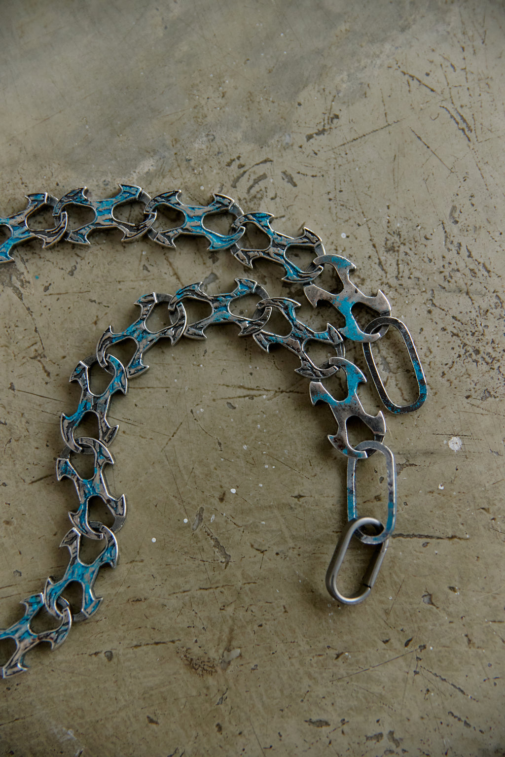 A close-up of a handmade silver-toned chain necklace with distressed turquoise blue enamel details, resting on a textured concrete surface.