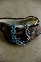 A close-up of a patterned zippered pouch or clutch with a distinctive turquoise and silver bicycle chain strap, resting on a textured concrete surface.