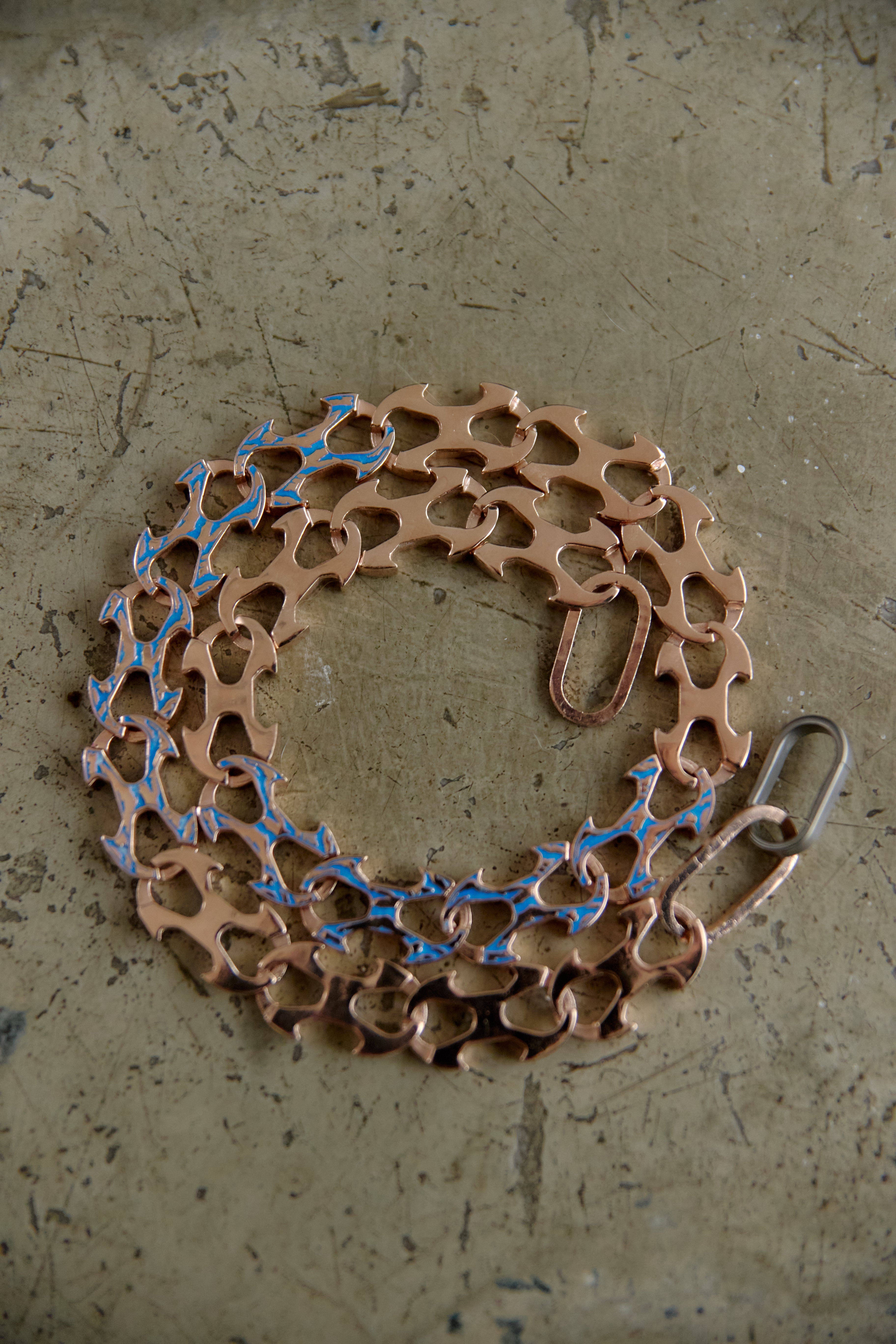 A bicycle cassette chain with copper-colored cogs and blue painted highlights, shown coiled on a textured concrete surface.