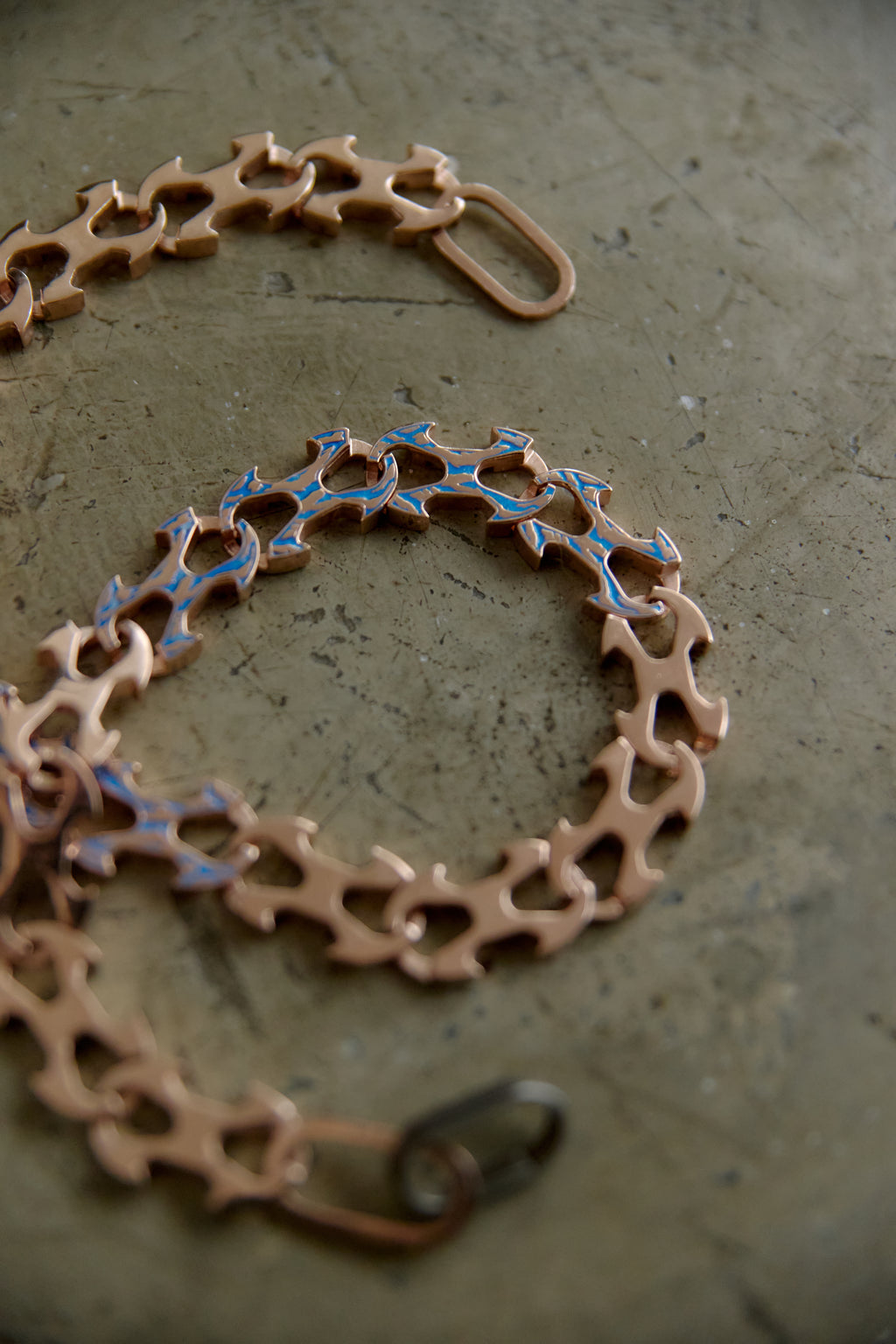 A close-up of a copper-toned chain bracelet with interlocking gear-like links and small blue inlay details, photographed on a textured gray surface.