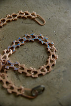 A close-up of a copper-toned chain bracelet with interlocking gear-like links and small blue inlay details, photographed on a textured gray surface.