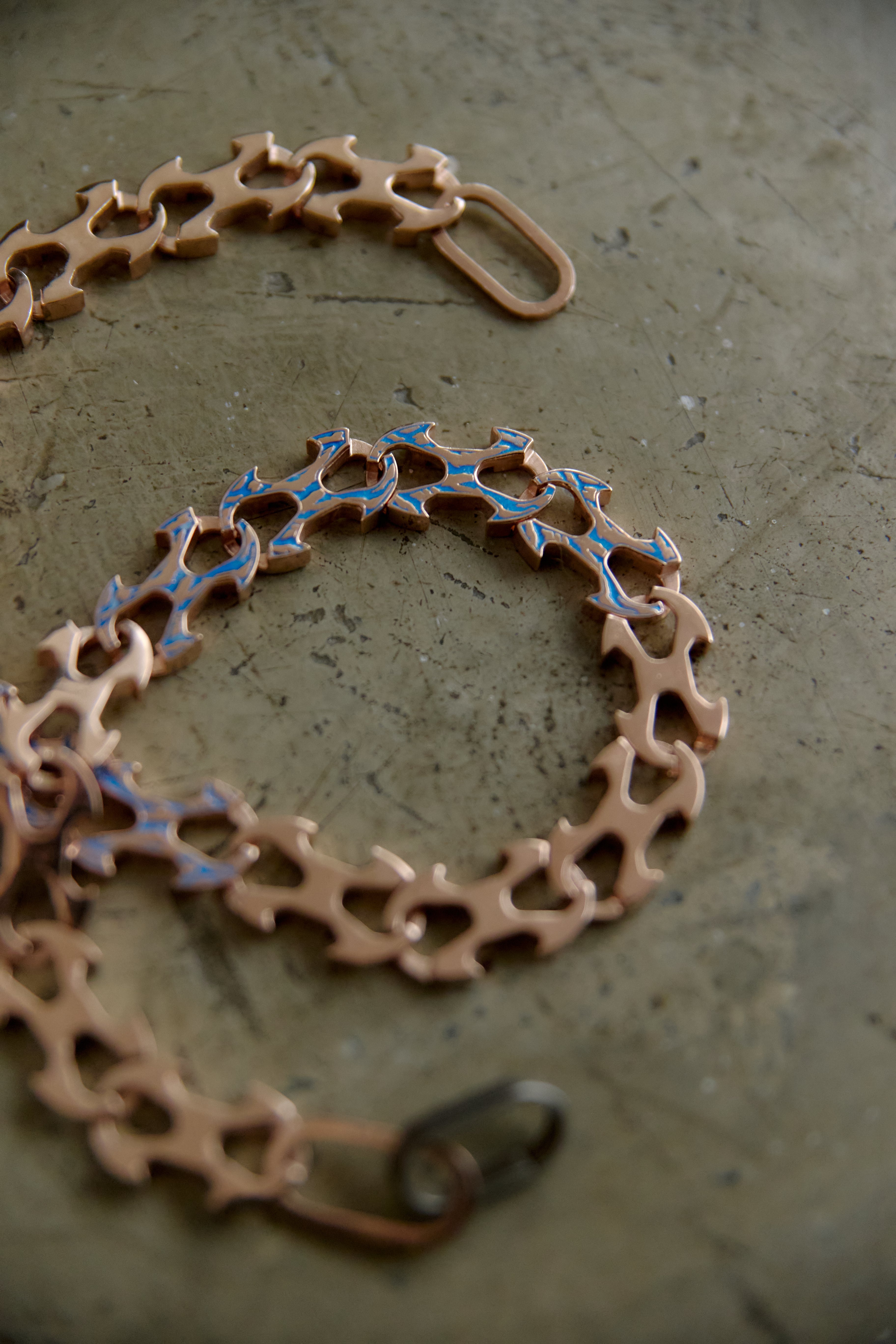 A close-up of a copper-toned chain bracelet with interlocking gear-like links and small blue inlay details, photographed on a textured gray surface.