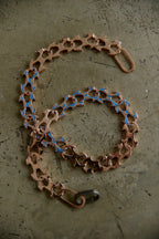 A bicycle chain-link style accessory with a copper-toned metal body and blue enamel accents, arranged on a textured gray surface.