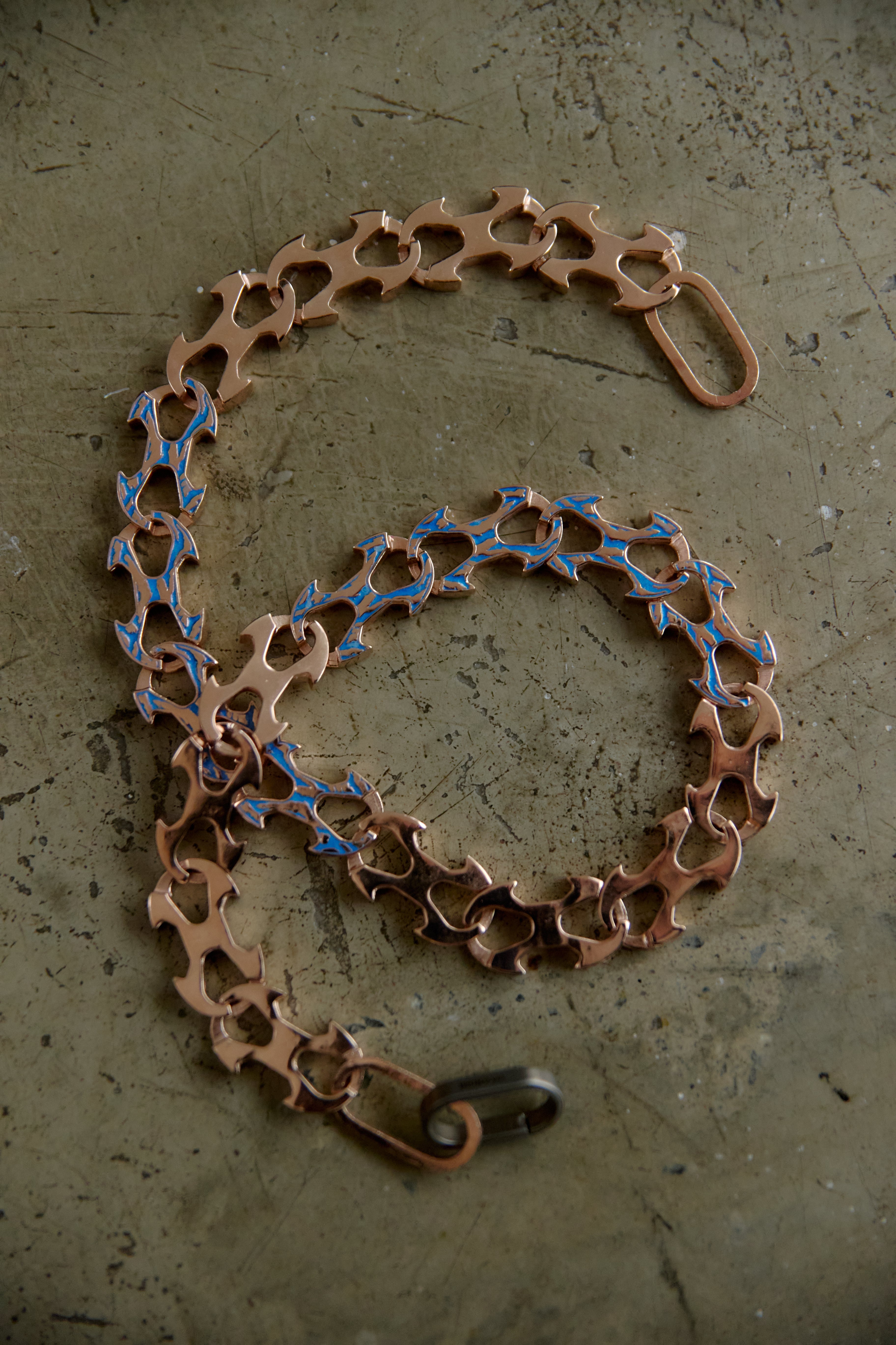 A bicycle chain-link style accessory with a copper-toned metal body and blue enamel accents, arranged on a textured gray surface.
