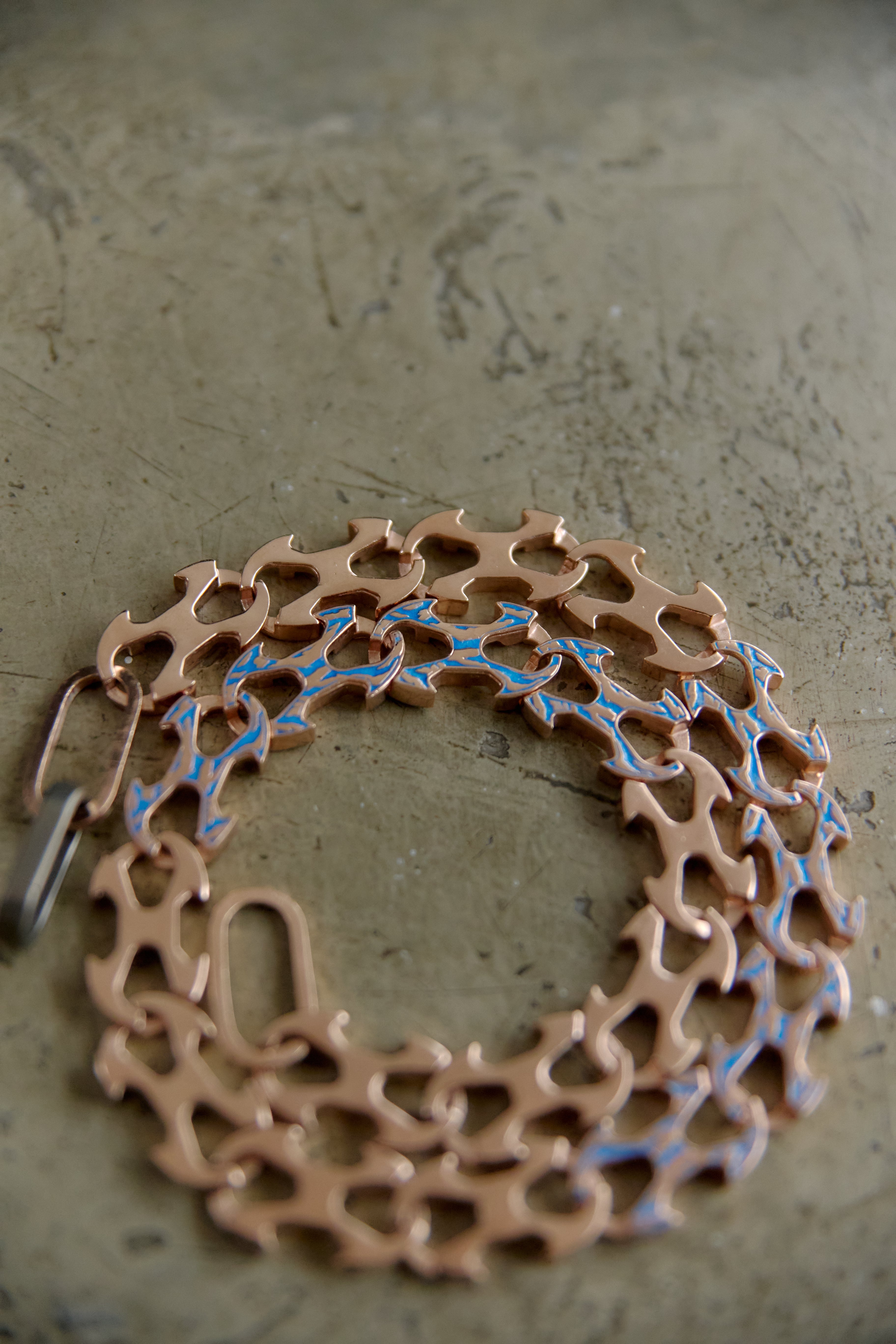 A copper-colored metal link chain bracelet with blue enamel accents, displayed on a textured gray surface.