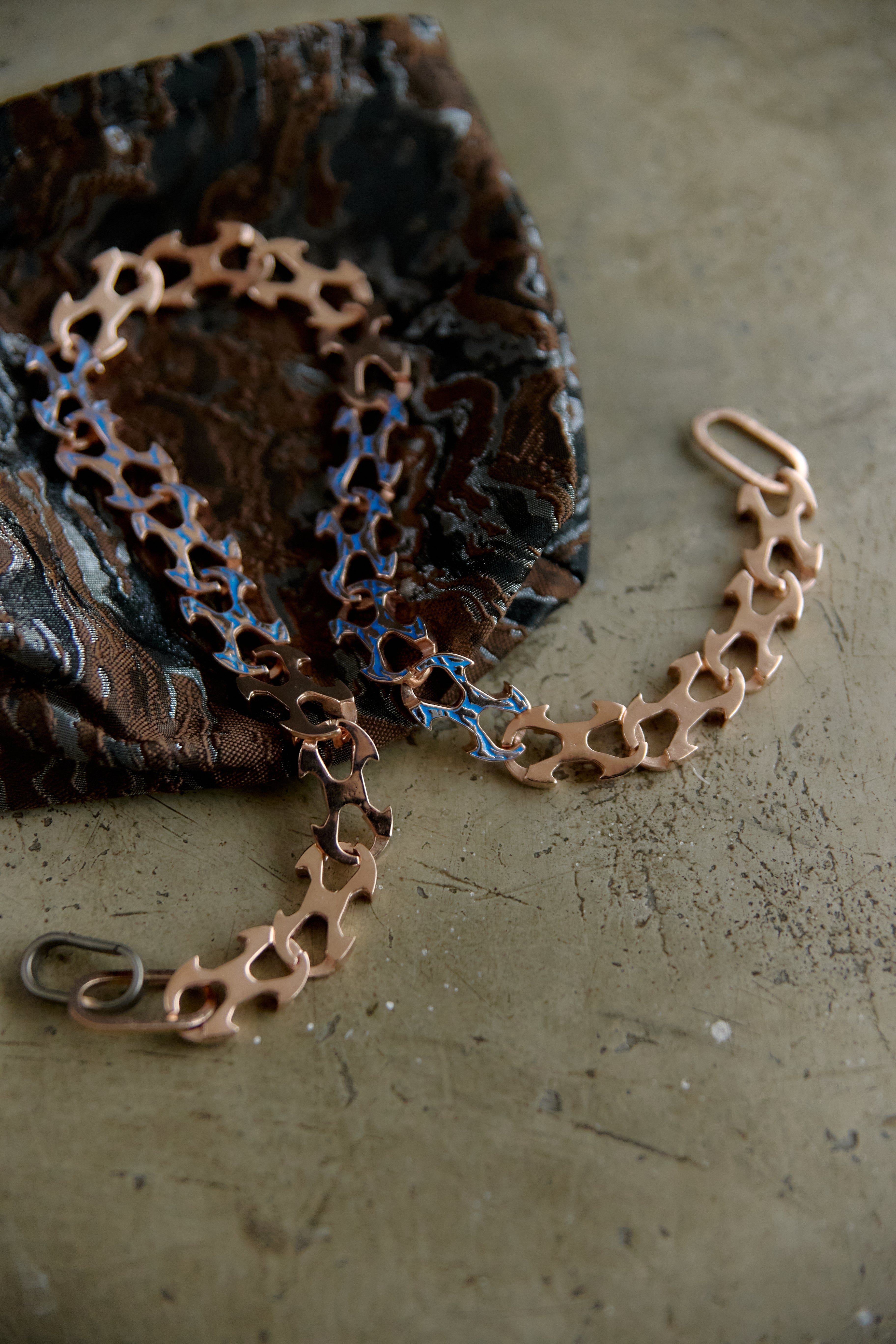 A copper-toned chain bracelet with a unique interlocking link design, draped over a dark patterned fabric on a textured beige surface.