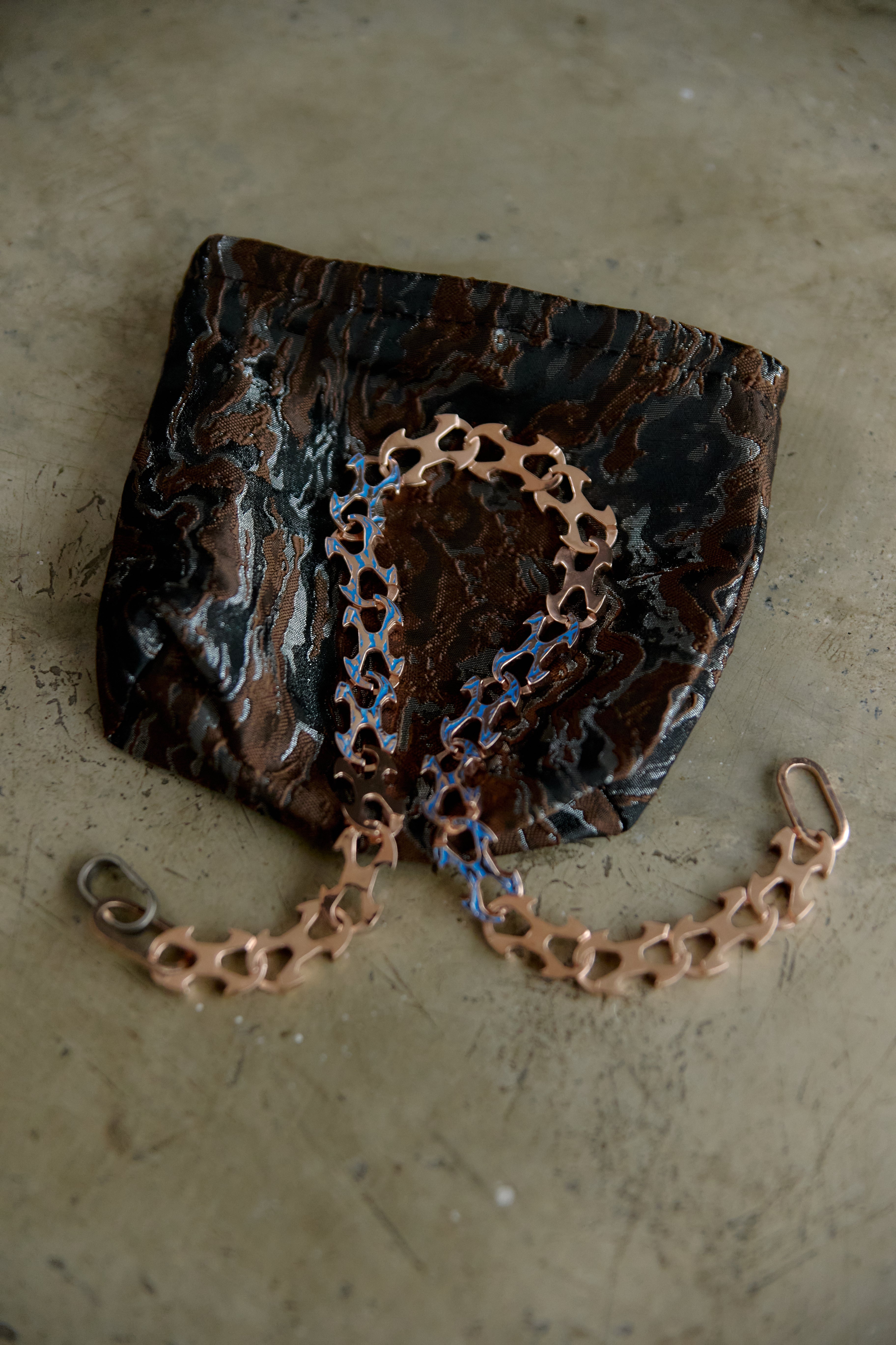A small black and brown marbled clutch purse with a copper heart-link chain strap, resting on a textured beige concrete surface.