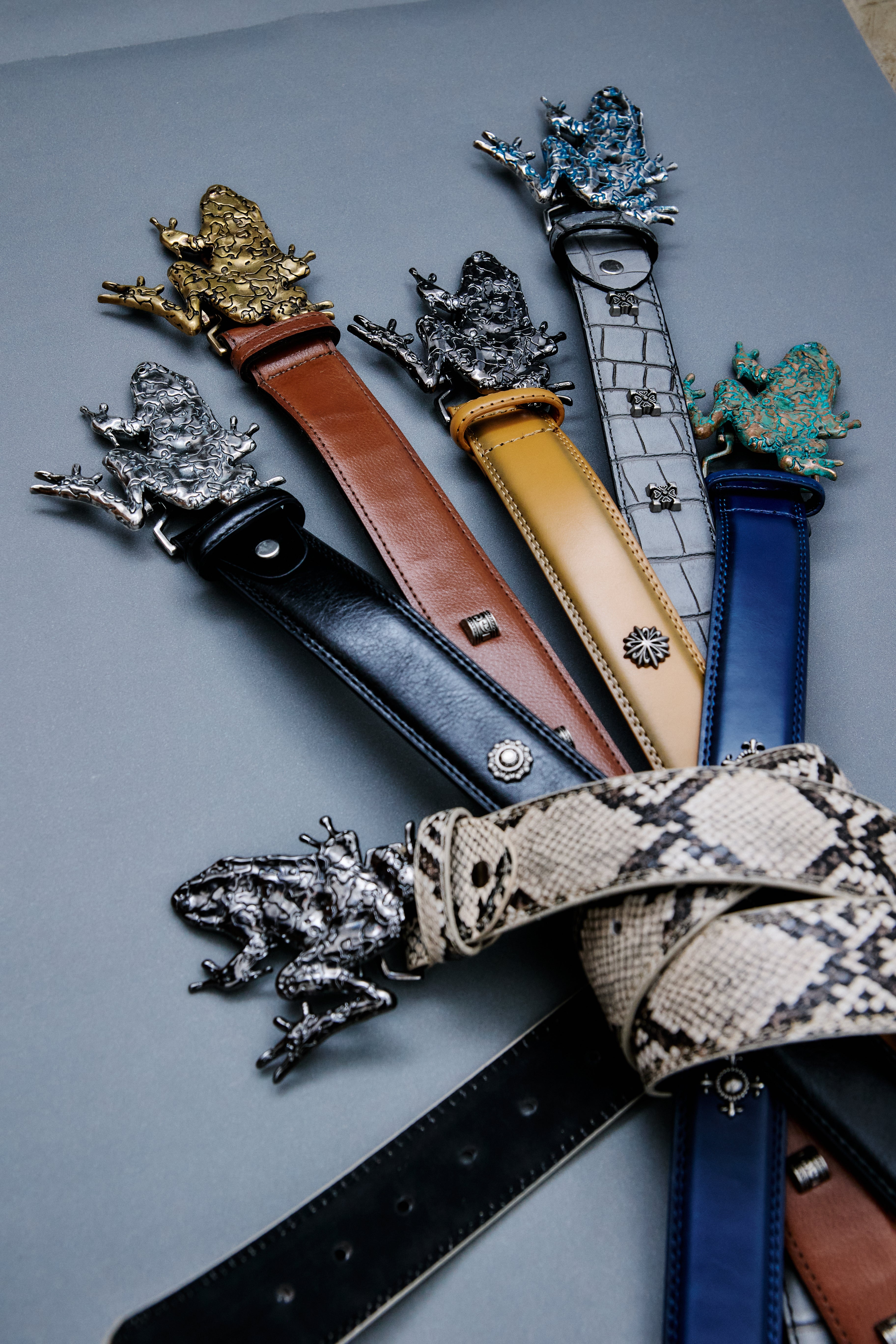 A set of fashion belts with detailed frog-shaped metal buckles, arranged on a smooth gray surface. The belts feature various materials and colors including black leather, brown leather, tan leather, white alligator print, blue leather, beige snakeskin print, and silver-tone metal.