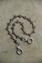 A handmade chain accessory featuring textured, scale-like links in brown, bronze, and blue, with silver-toned lobster clasps and hanging rings, displayed on a textured concrete surface.
