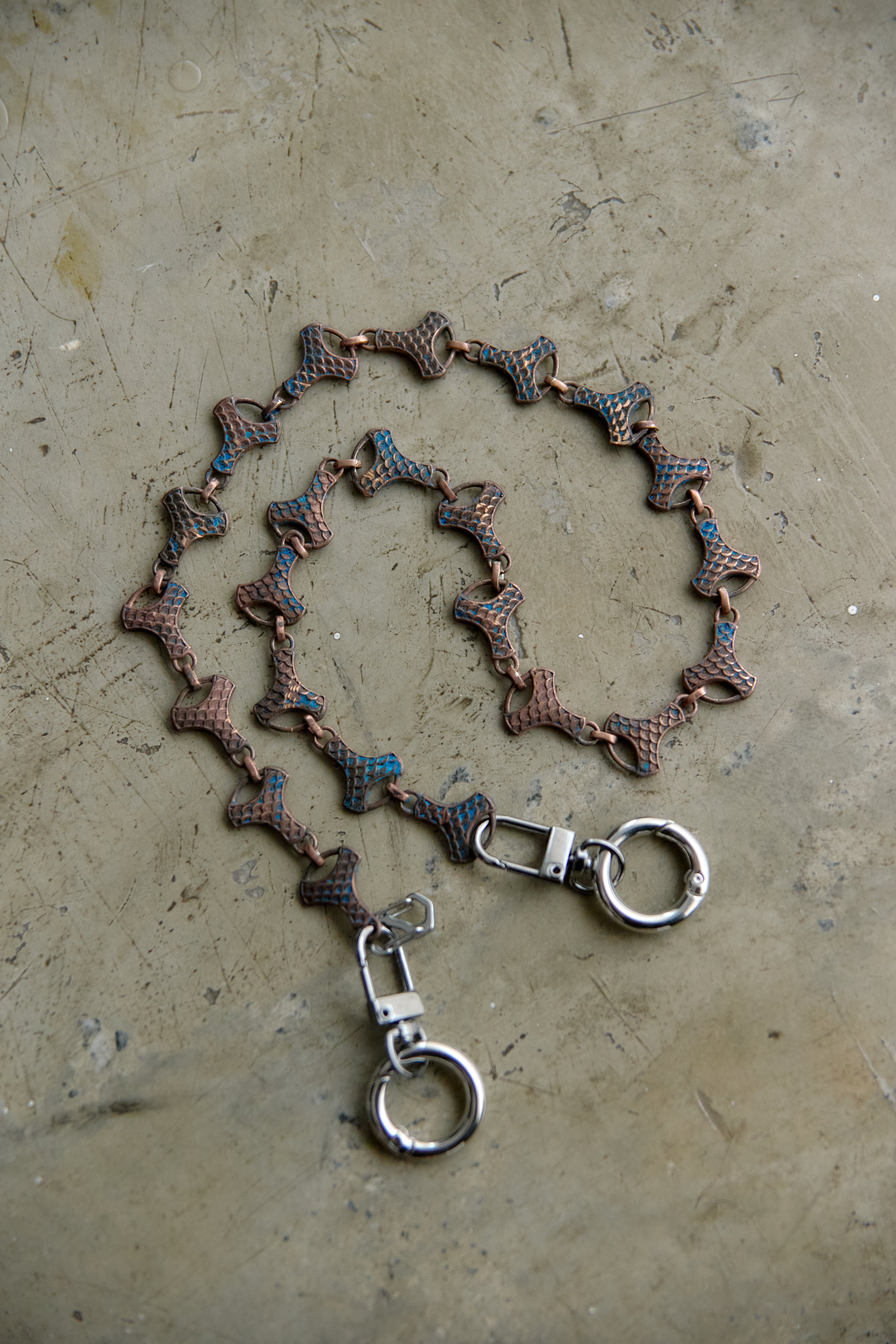 A handmade chain accessory featuring textured, scale-like links in brown, bronze, and blue, with silver-toned lobster clasps and hanging rings, displayed on a textured concrete surface.