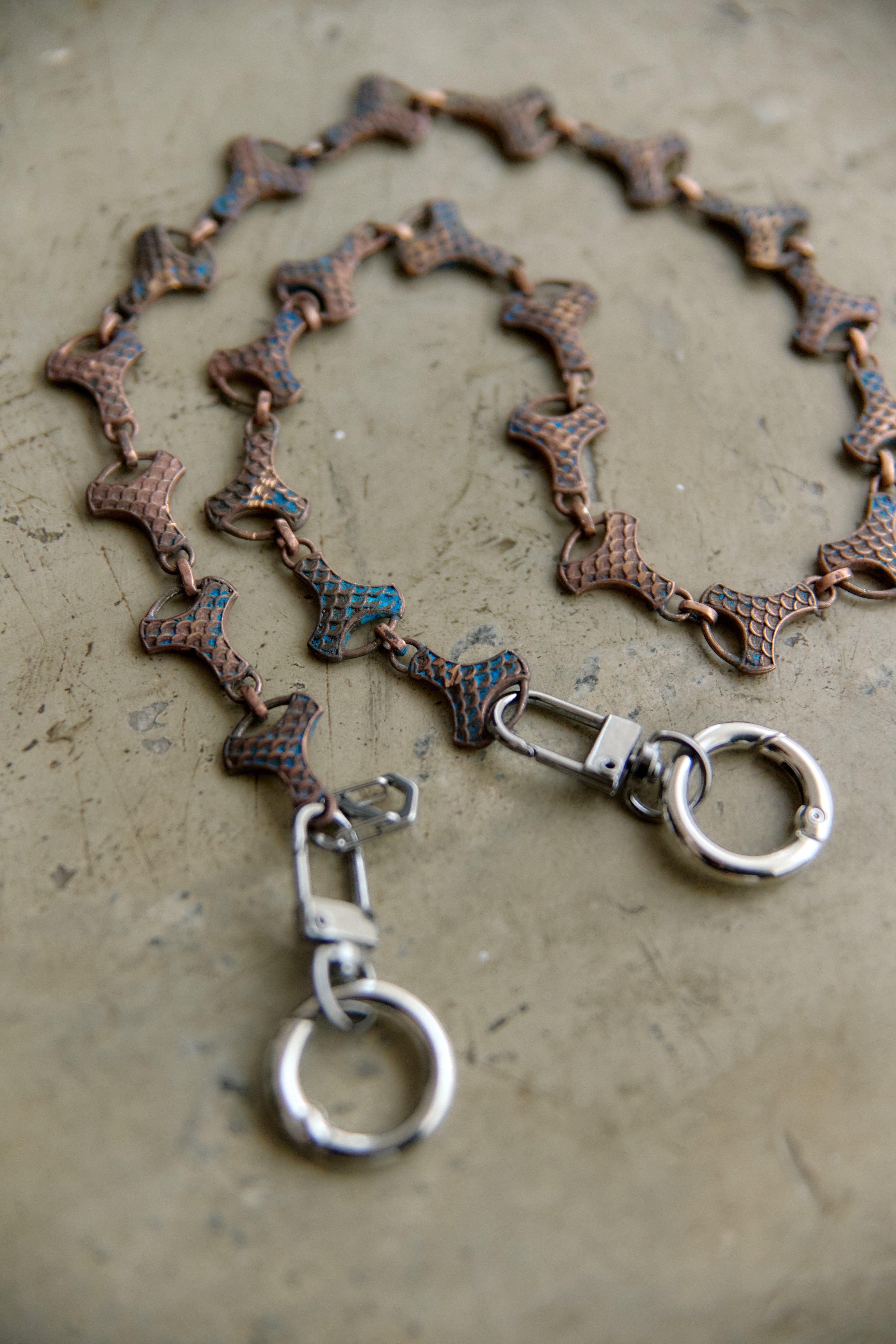 A close-up of a chain accessory with textured, metallic links in copper, bronze, and dark blue tones, featuring silver-toned lobster clasps and circular rings, laid on a textured gray surface.
