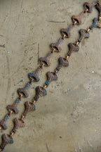 A close-up of a copper-toned chain necklace with textured, snake-like links featuring blue and gold enamel accents, laid diagonally on a textured beige surface.