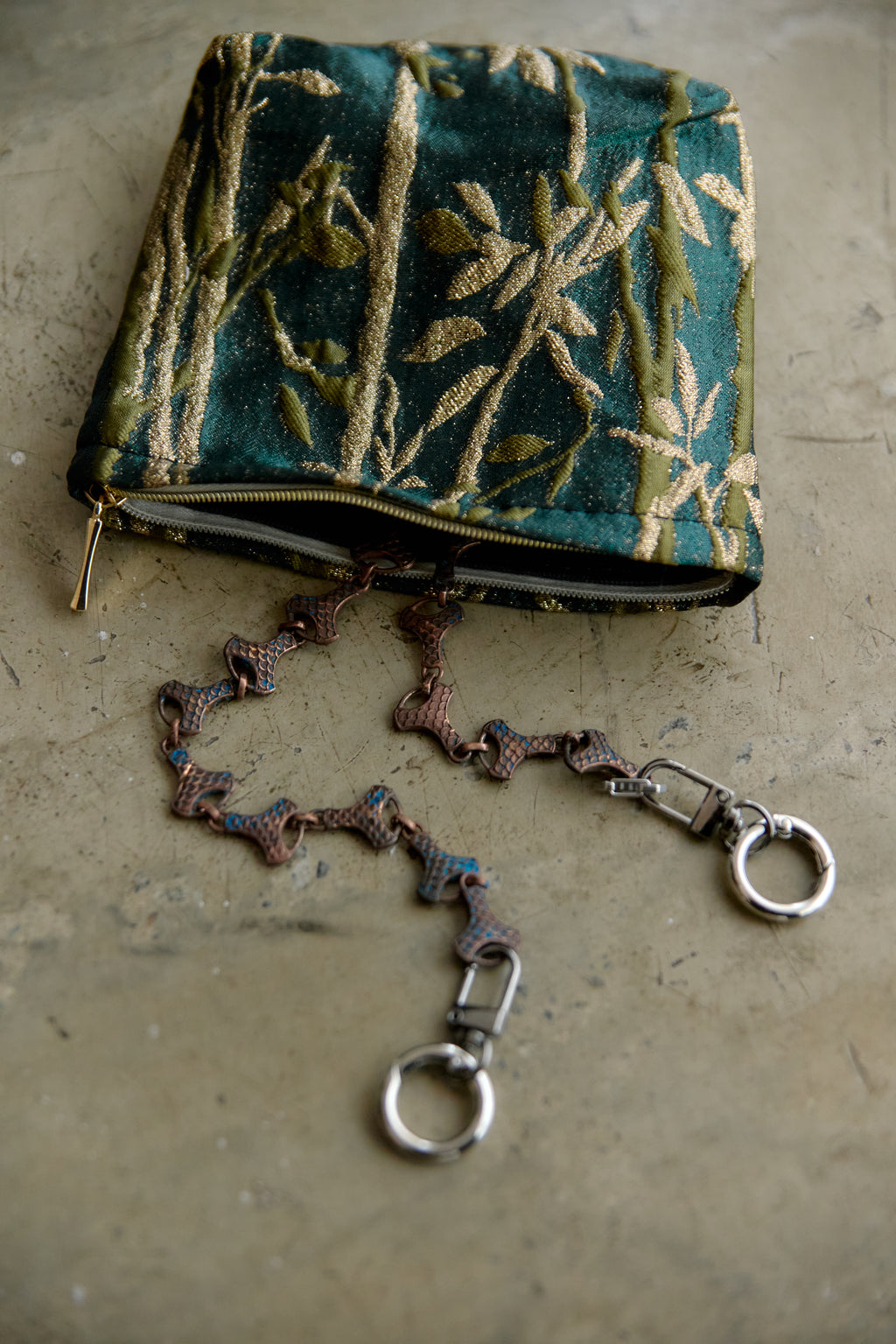 A small zippered clutch bag made of teal-green brocade fabric with gold floral patterns, featuring a detachable braided strap with silver-tone hardware, laid on a textured beige surface.