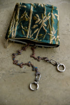 A small zippered clutch bag made of teal-green brocade fabric with gold floral patterns, featuring a detachable braided strap with silver-tone hardware, laid on a textured beige surface.