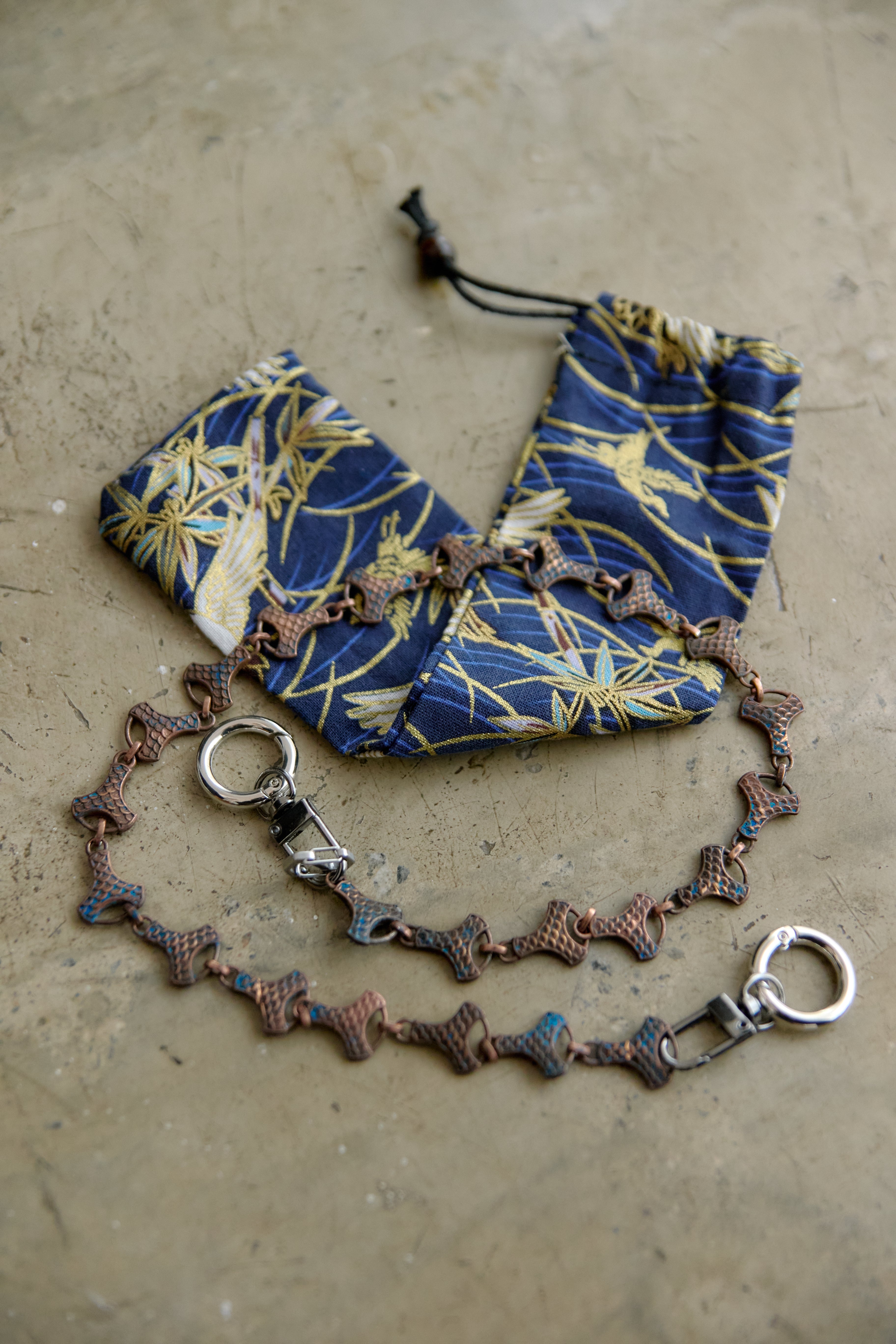 A handmade beaded chain accessory with metallic copper-toned links and silver-toned hardware, displayed next to a matching blue and gold patterned fabric pouch on a textured beige surface.