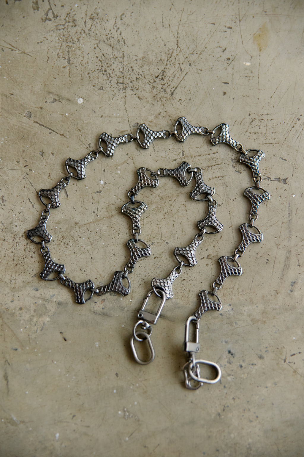 A graphite-toned metal chain accessory with repeating bow-shaped links and two lobster clasp ends, laid on a textured beige surface.