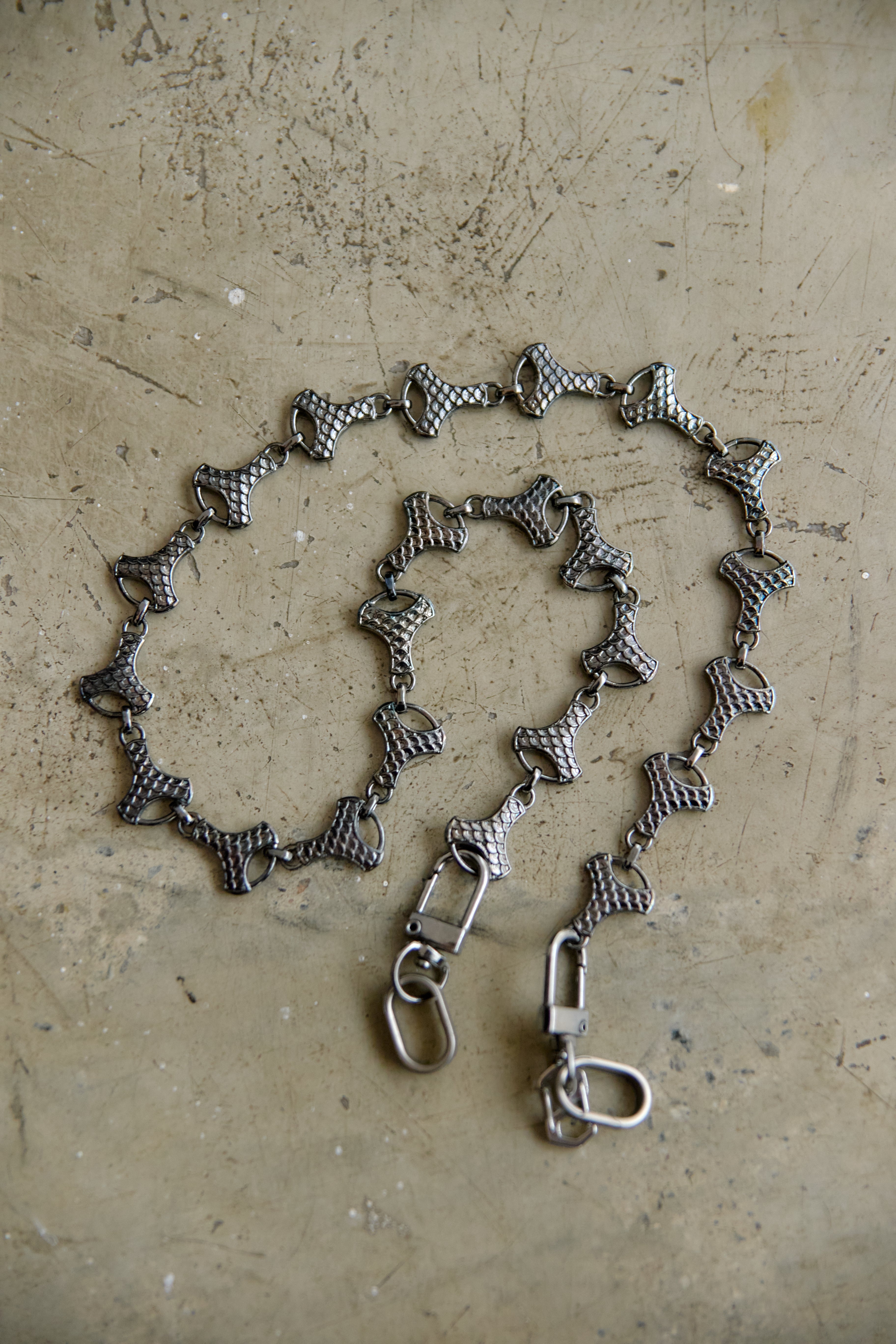 A graphite-toned metal chain accessory with repeating bow-shaped links and two lobster clasp ends, laid on a textured beige surface.