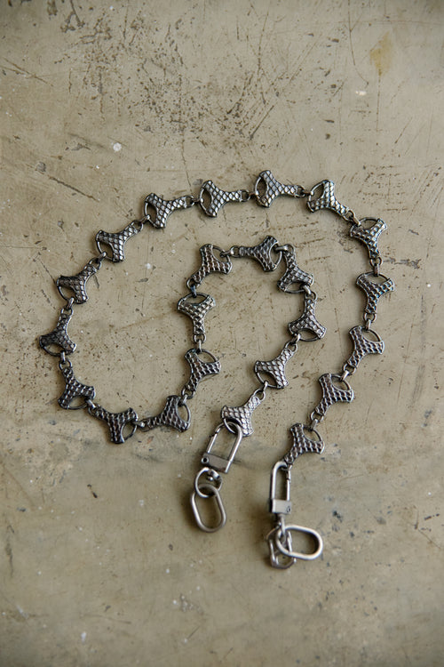 A graphite-toned metal chain accessory with repeating bow-shaped links and two lobster clasp ends, laid on a textured beige surface.
