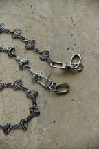 A close-up of a graphite-toned metal chain with repeating textured links and swivel snap clasps, laid on a mottled beige surface.