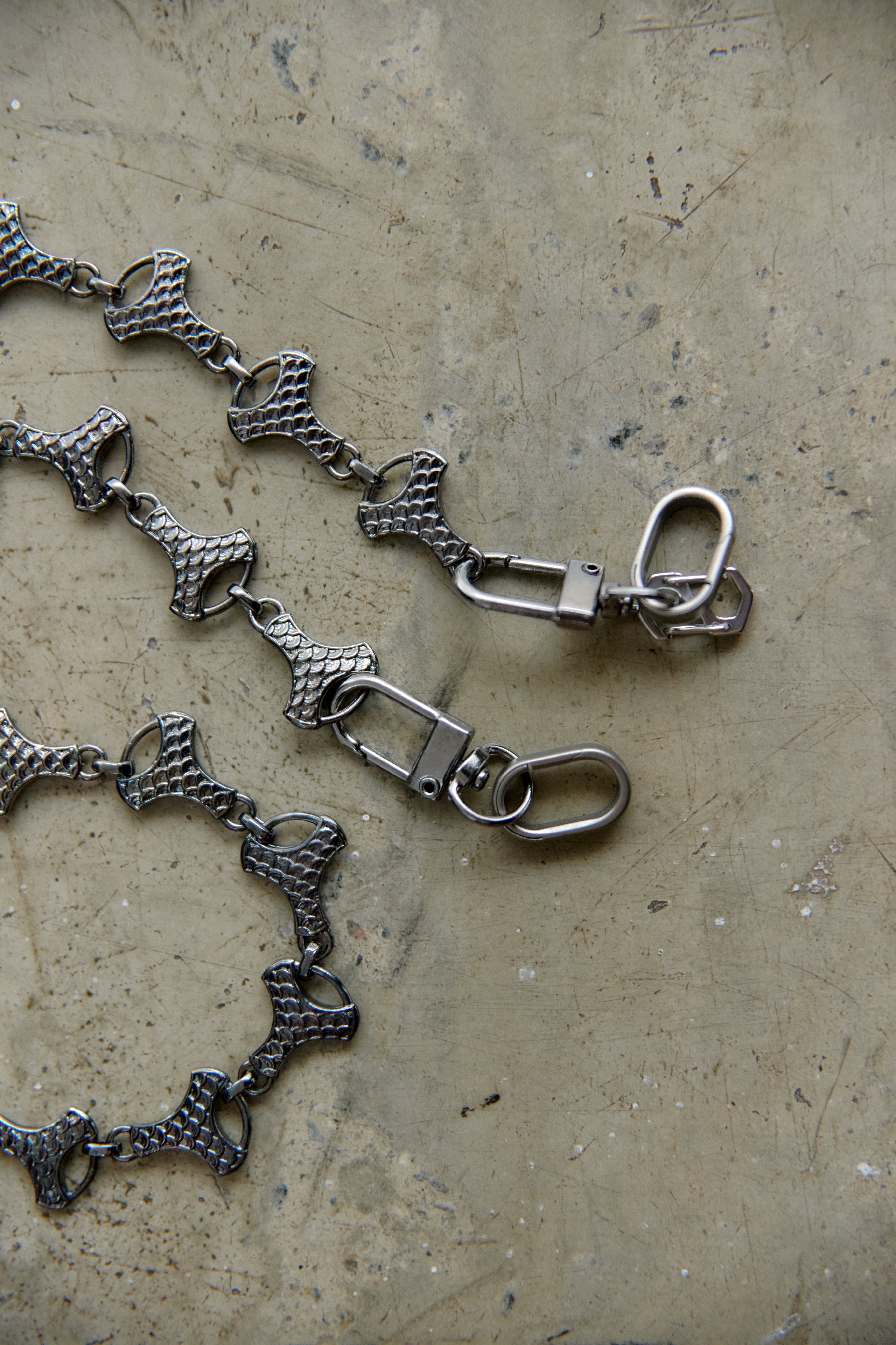 A close-up of a graphite-toned metal chain with repeating textured links and swivel snap clasps, laid on a mottled beige surface.