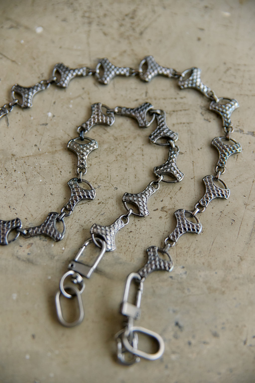 A graphite-toned metal chain necklace with textured, snake-like links and lobster clasps, displayed on a textured beige surface.