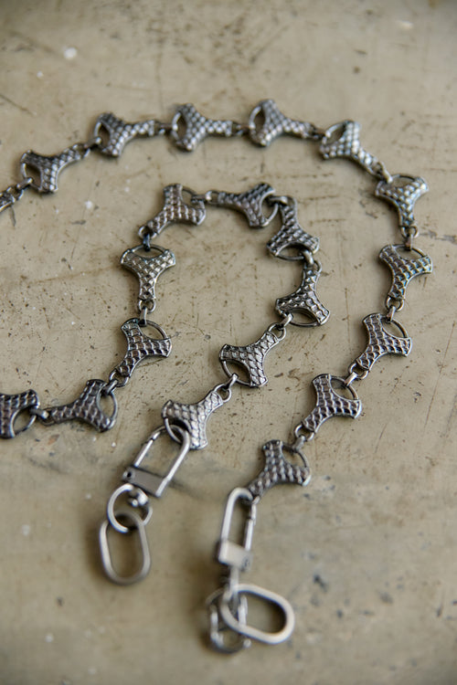 A graphite-toned metal chain necklace with textured, snake-like links and lobster clasps, displayed on a textured beige surface.