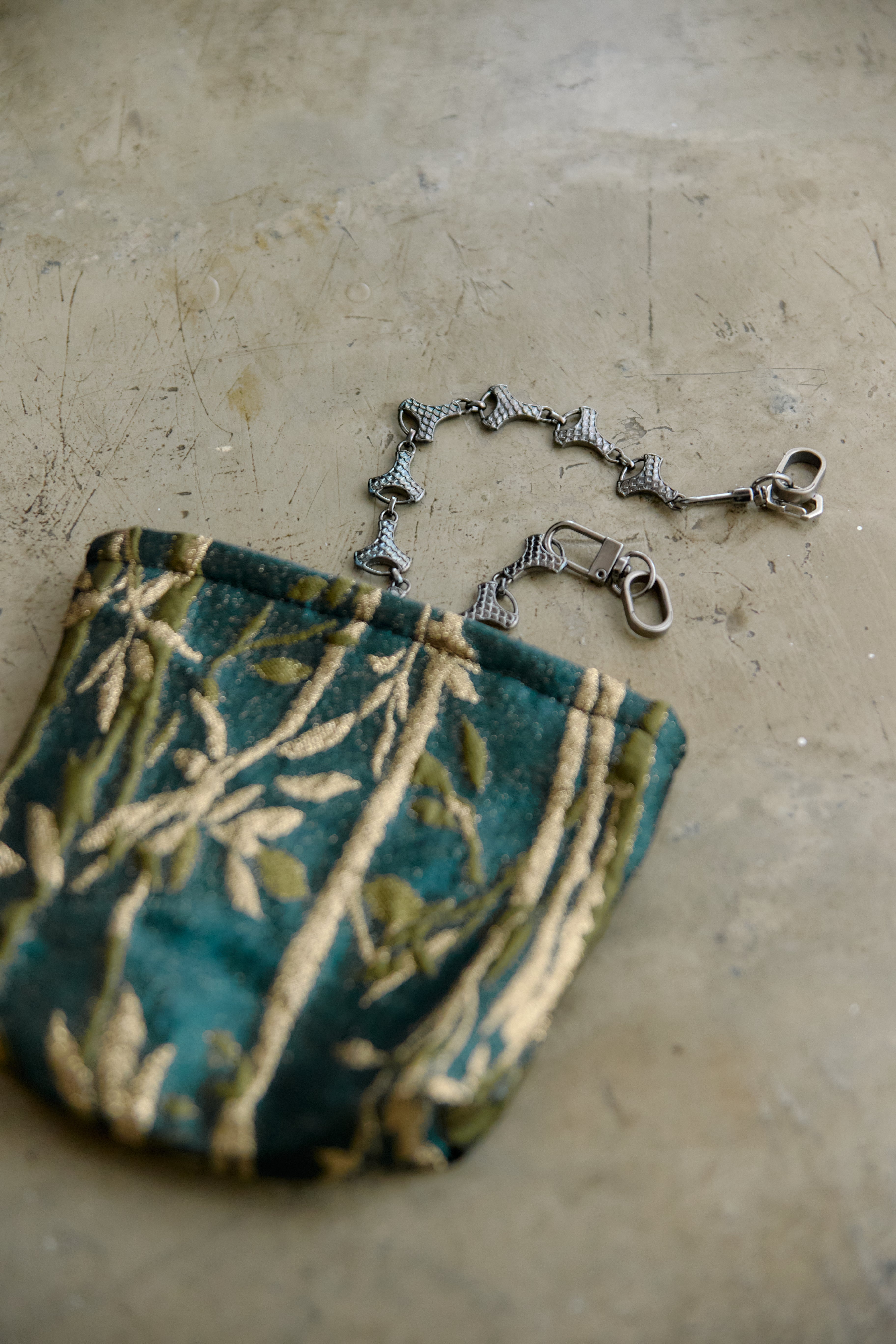 A small fabric pouch with a teal and olive green floral pattern, featuring a graphite metal chain strap with a lobster clasp, resting on a textured concrete surface.