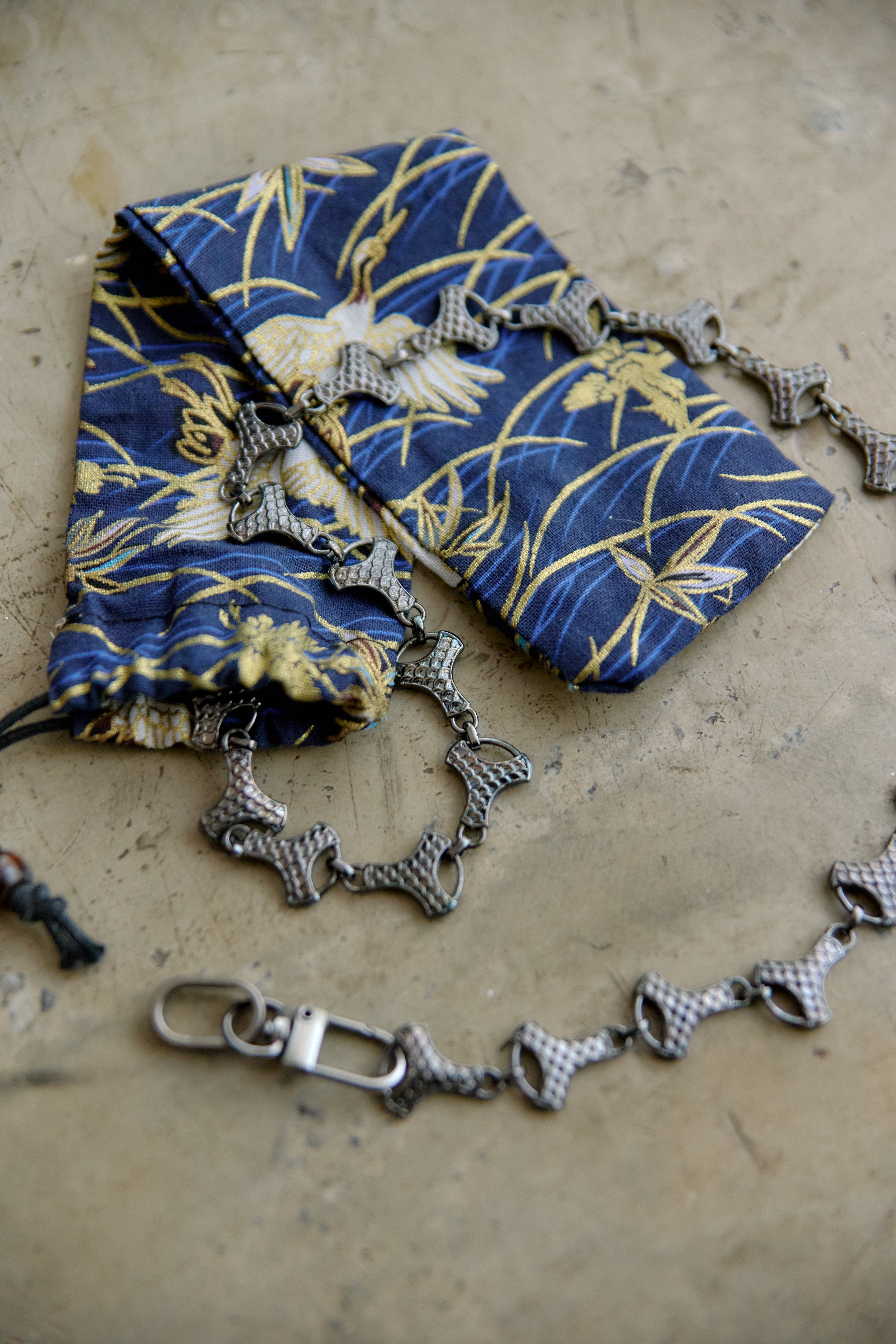 A graphite chain accessory with repeating bow-shaped links and a clasp, displayed on a beige textured surface next to a blue and gold patterned fabric pouch.