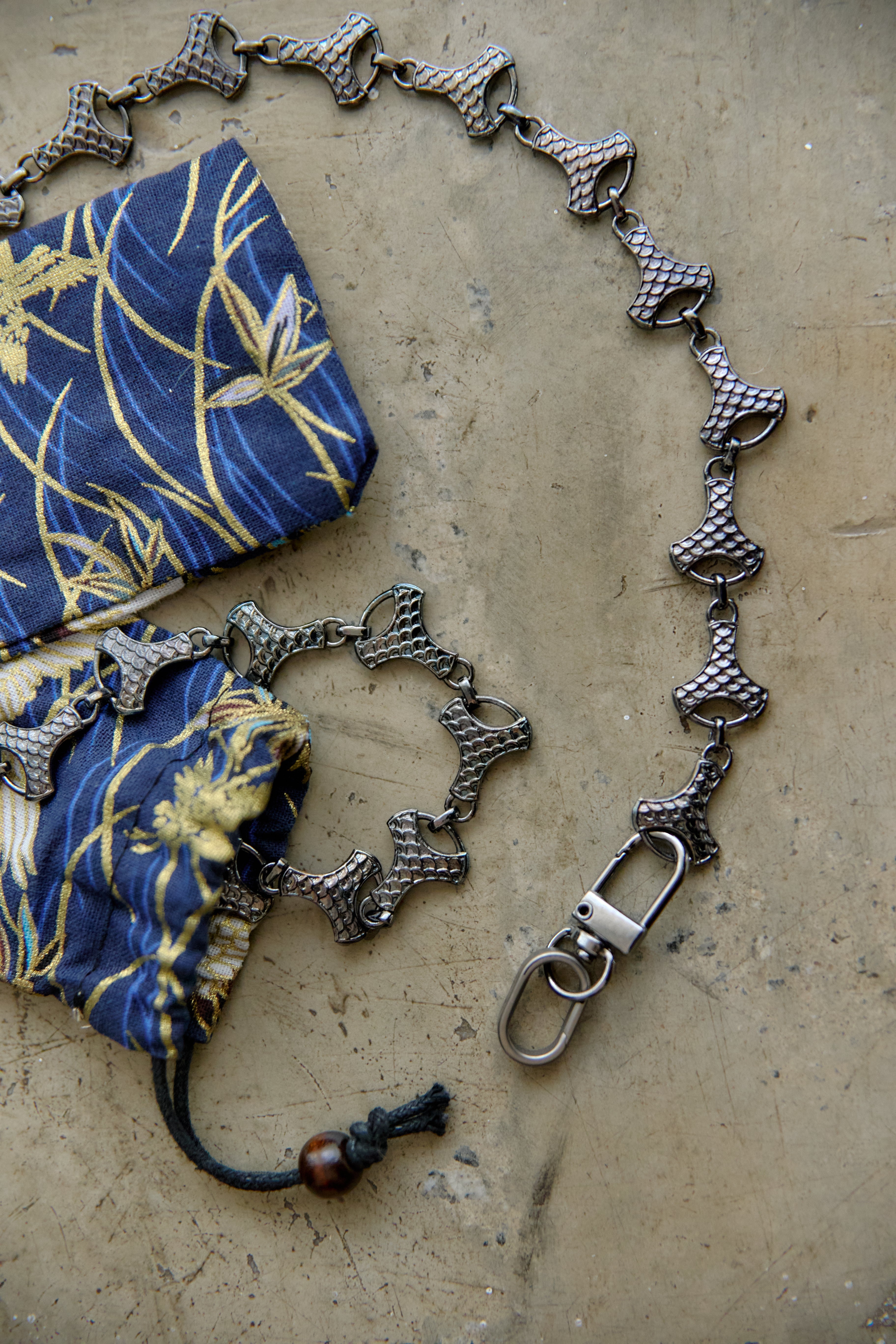 A graphite-toned metal chain accessory with textured bell-shaped links and a lobster clasp, displayed on a beige surface next to a blue and gold patterned fabric pouch.